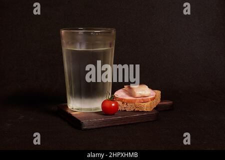 Bicchiere sfaccettato di vodka, panino con pane di segale, aringhe, prosciutto, pomodoro Foto Stock