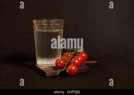 Un bicchiere sfaccettato di vodka e un rametto di pomodori ciliegini su un supporto di legno Foto Stock