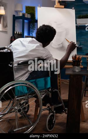 Studente d'arte in sedia a rotelle che disegna l'ilustrazione dei vasi lavorando all'ombra utilizzando strumenti grafici professionali durante la classe creativa. Il giovane illustratore che si ispira a tela in studio Foto Stock