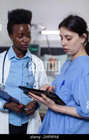 Infermiera che mostra i risultati di laboratorio da tablet digitale a medico che tiene appunti con storia medica nel reparto moderno dell'ospedale. Specialisti sanitari che utilizzano la tecnologia moderna per la diagnosi clinica. Foto Stock