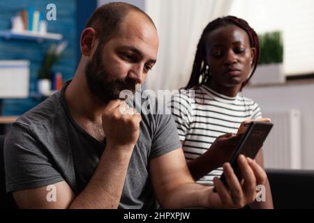 Uomo sicuro che prende selfie mentre annoiato la donna sta navigando in Internet e scorrendo sui social media utilizzando lo smartphone. Coppia multietnica seduta insieme sul divano in soggiorno mentre si utilizzano i moderni cellulari a casa Foto Stock