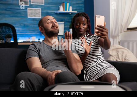 Felice coppia interrazziale sposata che sventolano al telefono moderno mentre in videocall. Allegra gente diversificata che parla con la propria famiglia attraverso una videocaletta online mentre si siede sul divano in salotto. Foto Stock