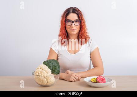 La donna caucasica preferisce cibo sano. Ragazza rossa sceglie tra broccoli e ciambelle su sfondo bianco. Foto Stock