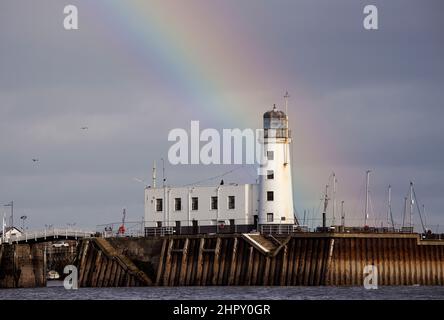 Un arcobaleno dona un tocco di colore a un cielo grigio invernale sul Mare del Nord. Il faro di Scarborough è un punto di riferimento locale per turisti e marittimi Foto Stock