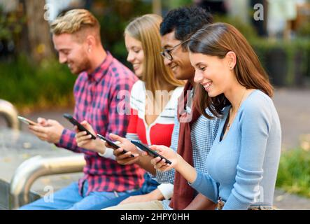 Gruppo di amici multirazziale positivi messaggi di testo su cellulari moderni mentre si siede insieme su strada con alberi verdi della città Foto Stock