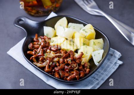 Funghi chanterelle con patate bollite, cipolle, aneto e olio in una piccola padella nera. Piatto tradizionale russo. Sfondo nero, primo piano, telecamera zo Foto Stock