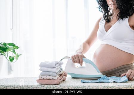 giovane donna piuttosto incinta stira i vestiti del suo prossimo bambino, preparando la stanza per il neonato Foto Stock