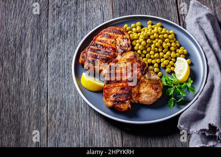 bistecche di carne di maiale alla griglia con piselli verdi bolliti e limone su un tavolo di legno, vista orizzontale dall'alto Foto Stock