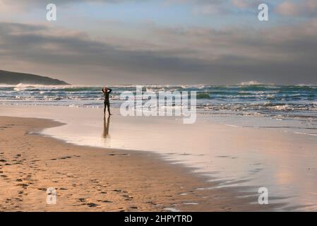 Un uomo che indossa un costume da bagno in piedi sulla riva e guarda verso il mare a Crantock Beach a Newquay in Cornovaglia. Foto Stock