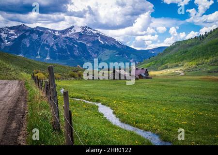 Alpine scene with dirt road rustic barn and snow runoff stream running through valley with wildflowers - snow peaked mountains near Crested Butte Colo Foto Stock