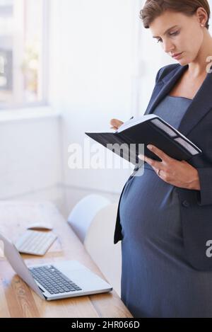 Confermando il suo congedo di maternità. Una donna d'affari incinta che legge il suo pianificatore quotidiano nel suo ufficio. Foto Stock