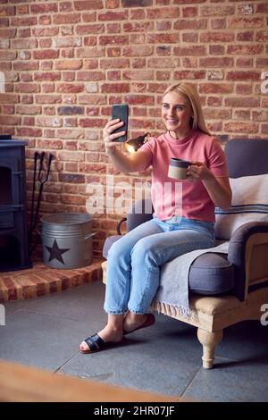 Giovane donna seduta in poltrona contro mattoni esposti prendendo Selfie al telefono Foto Stock