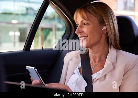 Donna in viaggio d'affari matura in taxi o in auto che indossa auricolari durante la chiamata in conferenza utilizzando il telefono cellulare Foto Stock