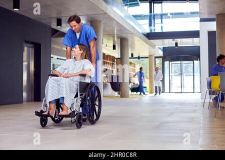Infermiera maschile che indossa scrub spingendo il paziente femminile in sedia a rotelle attraverso l'edificio dell'ospedale Foto Stock