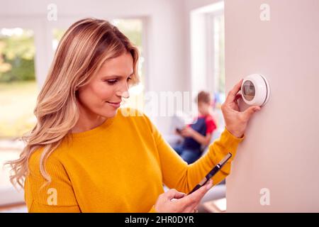 Madre a casa con Son Adjusting Smart Central Heating Thermostat Control con App su cellulare Foto Stock