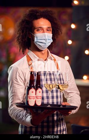 Ritratto di Server maschio indossando maschera viso vassoio di tenuta di bevande al Cool Bar o Club Foto Stock