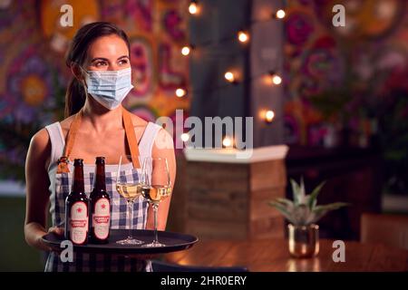 Ritratto di Server femmina indossando maschera viso vassoio di tenuta bevande al Cool Bar o Club Foto Stock