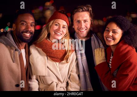 Ritratto di amici all'aperto indossando cappotti e sciarpe incontro contro le luci della città di notte Foto Stock