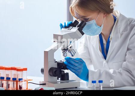 Lavoratore di laboratorio femminile che indossa i DPI per l'analisi dei campioni di sangue in laboratorio con microscopio Foto Stock