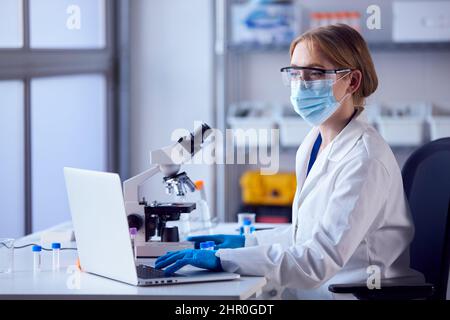 Camice da laboratorio femminile che indossa un camice da laboratorio per l'analisi dei campioni in laboratorio con notebook e microscopio Foto Stock
