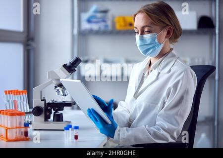 Camice da laboratorio femminile che indossa un camice da laboratorio che lavora sul vaccino Covid-19 con tavoletta digitale e microscopio Foto Stock