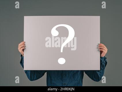 Un giovane tiene un grande foglio con un punto interrogativo, chiedendo perché, cercando risposte, sfondo scuro Foto Stock