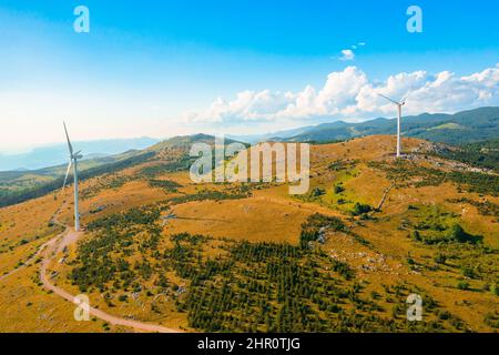 Le turbine eoliche generano energia pulita alternativa in piedi sulle colline con cespugli in altopiano contro le montagne forestali in vista aerea di giorno di sole Foto Stock