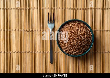 Il grano saraceno organico crudo groats in una ciotola e forchetta sopra il mat marrone della tabella di legno. Grani di grano saraceno arrosto. Concetti di cibo vegetariano, dieta senza glutine. Foto Stock