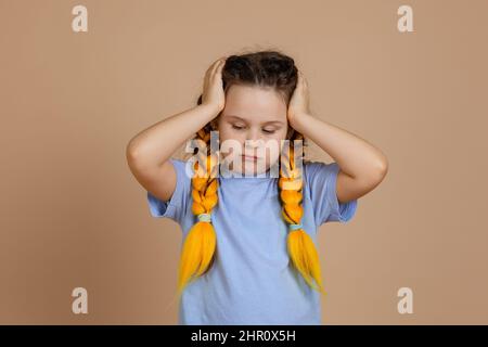 Arrabbiato caucasico bambino femmina che tiene testa con le mani guardando verso il basso avendo kanekalon trecce di colore giallo sulla testa in blu t-shirt sul beige Foto Stock