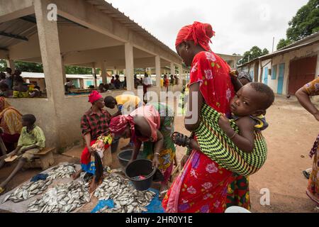 Una madre porta il suo bambino sulla schiena mentre lei negozi di pesce fresco in un mercato a Tanaff, Senegal, Africa occidentale. Foto Stock