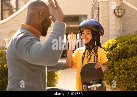 Sorridente ragazza afroamericana che indossa casco tenendo skateboard dando alto-cinque a padre in cantiere Foto Stock