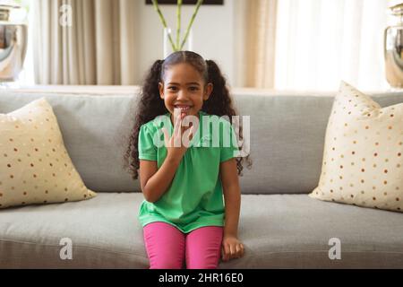 Ritratto di sorridente ragazza muta biraciale che parla con linguaggio del segno della mano seduto sul divano a casa Foto Stock