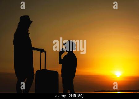 Madre e figlio giovane con una valigia sullo sfondo del tramonto del mare. Un viaggio al mare, una vacanza, l'oceano, un viaggio in paesi caldi. Foto Stock