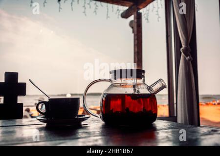 Tè in una teiera di vetro e una tazza su un tavolo di legno all'aperto. Caffè costiero, brunch. La spiaggia e il mare sono sfocati. Tè fresco, calmo. Foto Stock