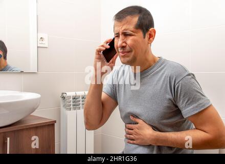 Scatto corto di un uomo seduto sul gabinetto in un bagno che soffre di crampi di stomaco e chiamare assistenza medica al telefono Foto Stock