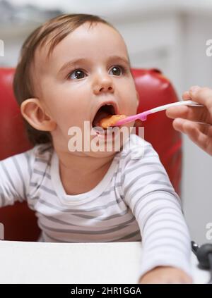 Apertura ampia.... Scatto di un bambino piccolo carino seduto in un seggiolone ed essendo alimentato dalla sua mamma. Foto Stock