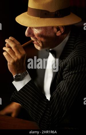Si accende. Vecchio boss mob che indossa un cappello e guarda serio mentre si accende una sigaretta. Foto Stock