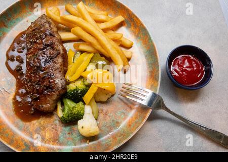 Bistecca alla griglia con salsa, patatine fritte e verdure assortite su un piatto accanto al ketchup in un contenitore monouso per ketchup Foto Stock