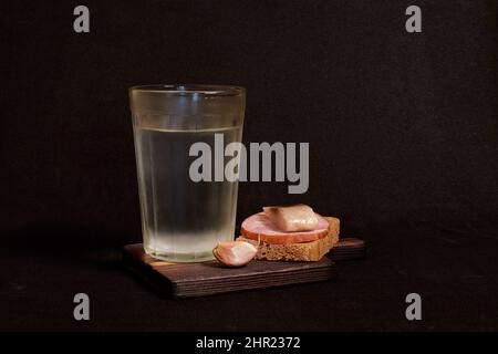 Bicchiere sfaccettato di vodka, panino con pane di segale, aringhe, prosciutto, spicchi d'aglio Foto Stock