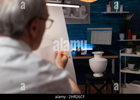 Focus sul modello di vaso bianco in gesso utilizzato per lo studio della luce disegnato da un uomo anziano in studio d'arte. Dettaglio di un oggetto fermo e di un artista in pensione che disegnano un disegno su carta utilizzando la matita in un laboratorio creativo. Foto Stock