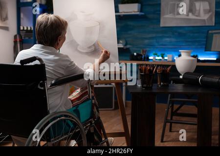 Messo a fuoco insegnante di arte pensionato che vive con disabilità facendo arte creativa in matita disegno modello vaso su carta tela su cavalletto. Donna più anziana che utilizza uno schizzo ombreggiato ispirato alla sensazione di sedia a rotelle. Foto Stock