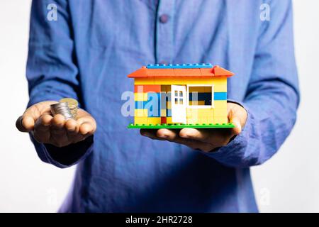 Fuoco selettivo sulle monete e sulla casa del giocattolo tenuto in mano da un uomo su sfondo bianco Foto Stock