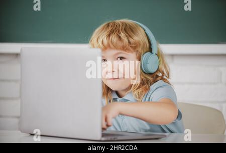 Primo piano ritratto di bambino piccolo in cuffie guardare video lezione sul computer a scuola, bambino in cuffie hanno lezione online, utilizzando il computer portatile. Foto Stock