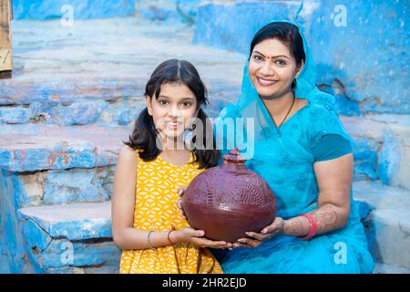 Ritratto di madre indiana felice e figlia che tiene in mano scatola di denaro di argilla o gullak. Donna sorridente che indossa sari e ragazza con la tradizionale banca di maialino Foto Stock