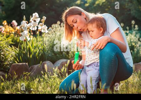 Buon giorno per i bambini. La giovane madre abbraccia la figlia del bambino e soffia le bolle di sapone. Giochi estivi con un bambino nel cortile. Parco soleggiato sul retro Foto Stock