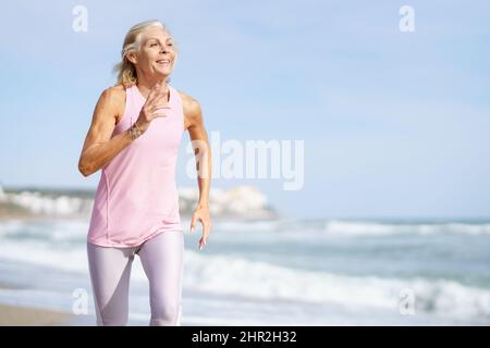 Donna matura che corre lungo la riva della spiaggia. Donna più anziana che fa sport per mantenere la misura Foto Stock