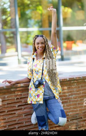 Donna nera con trecce africane, sollevando il braccio con gioia. Ragazza che tiene una macchina fotografica. Foto Stock