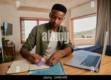 Giovane adulto nero maschile scrittura nota su adesivo accanto al portatile, concentrando. Seduta in appartamento moderno che lavora da casa. Foto Stock