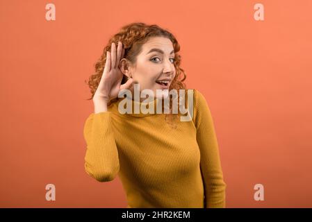 Giovane e attraente ragazza rossa caucasica che tiene la mano vicino al suo orecchio a causa della sordità isolato su sfondo arancione studio. Foto Stock