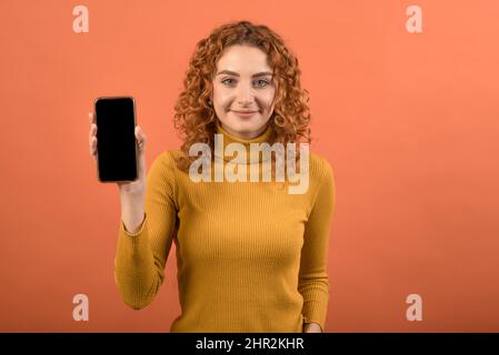 Giovane e attraente ragazza rossa caucasica in arancione jumper tenere smartphone, mostrando applicazione, isolato su sfondo arancione studio. Foto Stock